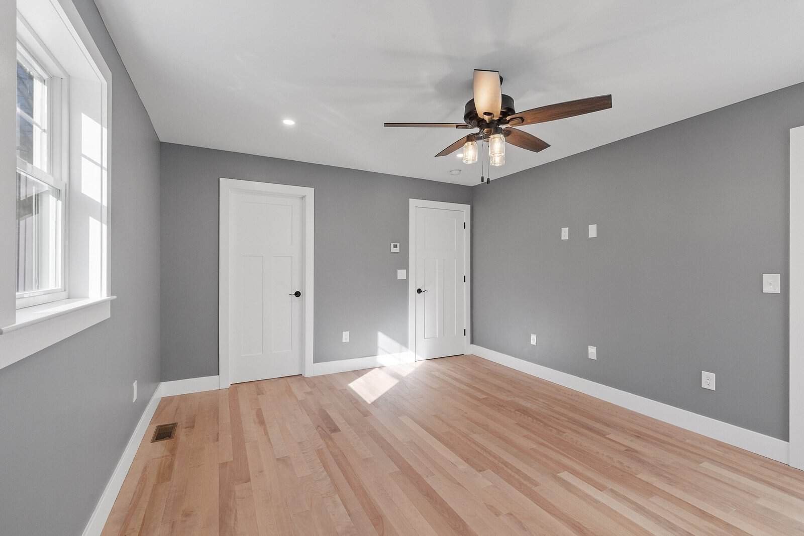 Empty room with gray walls, hardwood floor, ceiling fan, and natural light from windows.